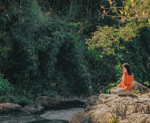 Woman meditating by a serene riverside surrounded by lush greenery on a sunny day.